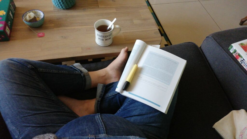 Une femme enceinte assise en tailleur lisant un livre avec une tasse de thé.