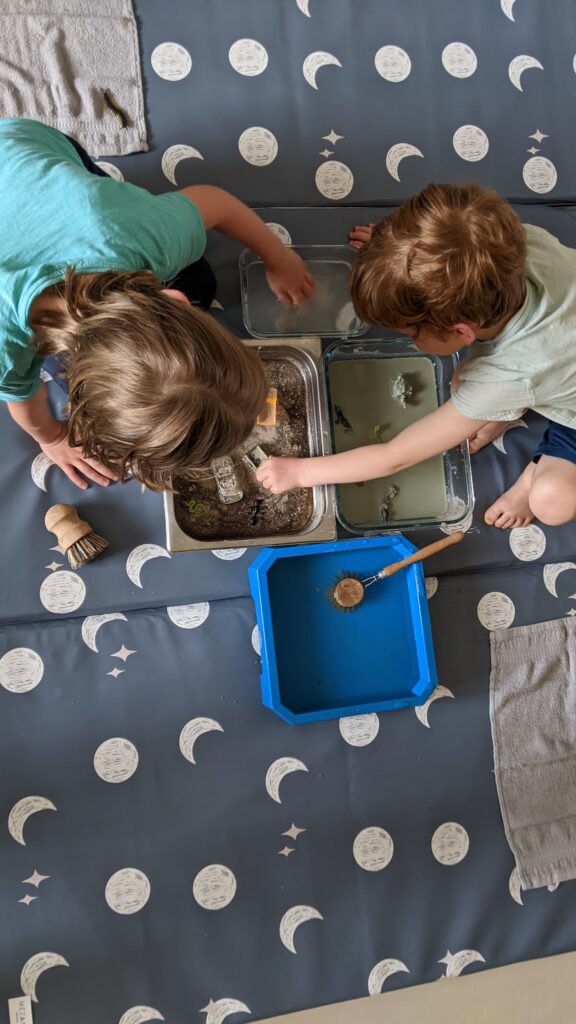 Photo de deux enfants faisant une activité salissante sur un tapis : ils nettoient des figurines à l'aide d'une brosse.