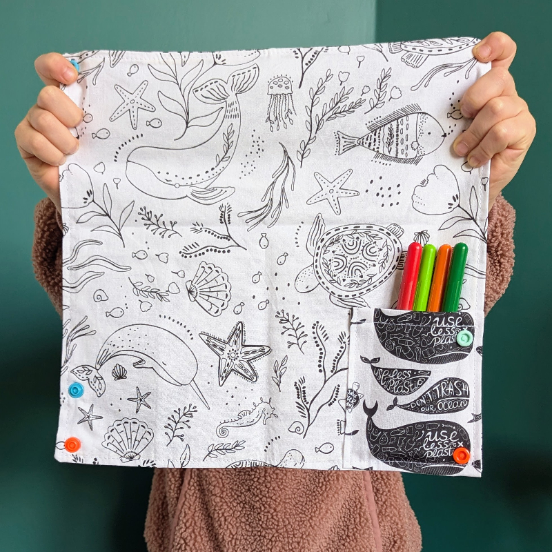 Enfant qui tient en main un coloriage lavable nomade de la sardine verte sur le thème des animaux marins. Quatre feutres à l'eau sont rangés dans la pochette cousue au cahier.