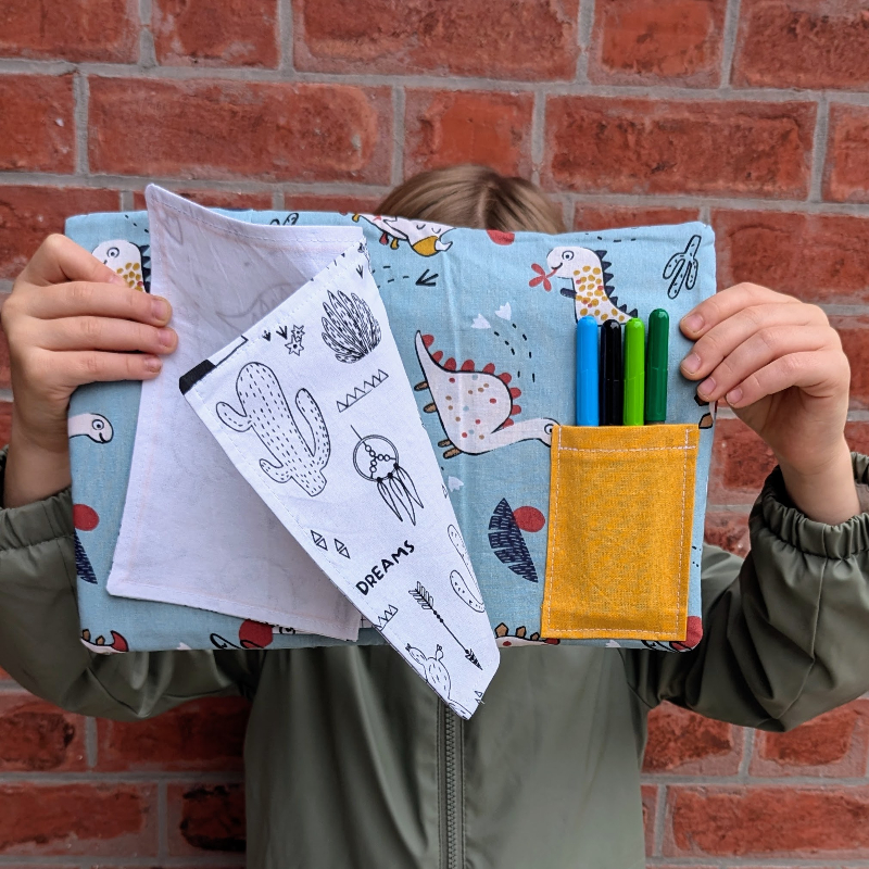 enfant tenant un cahier d'activités lavable de la sardine verte ouvert sur un coloriage de tipis et de cactus. On aperçoit aussi une pochette de feutres à l'eau cousue au cahier.