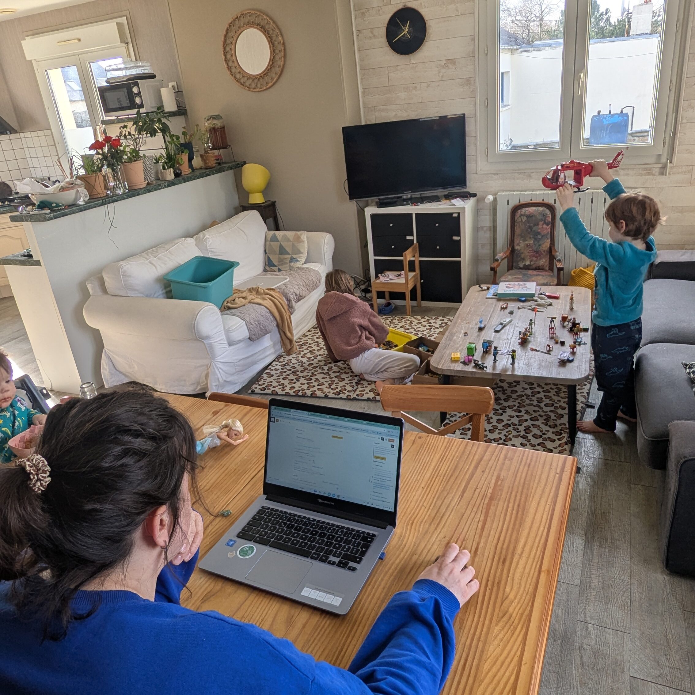 photo d'une femme travaillant sur ordinateur pour son nouveau blog, dans son salon avec deux enfants jouant aux playmobils, et un bébé qui mange dans sa chaise haute 